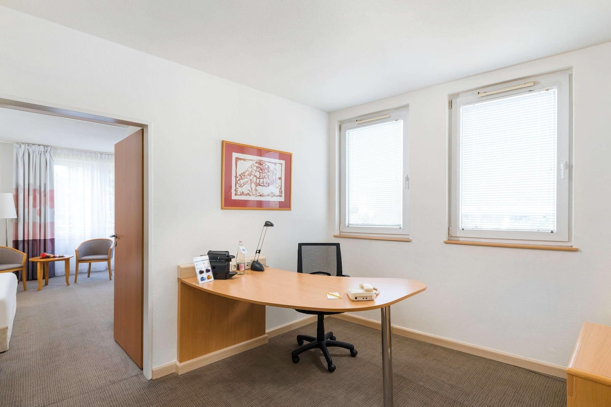 A hotel room with a desk, chair, and a bed. There are two windows with blinds, a door, and a framed picture on the wall.