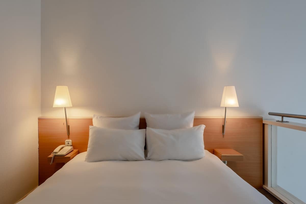 A hotel room with a bed, two bedside lamps, a nightstand with a phone, and a window.