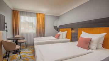 A hotel room with two beds, a small table, chairs, a mirror, and a view of the city through the window.