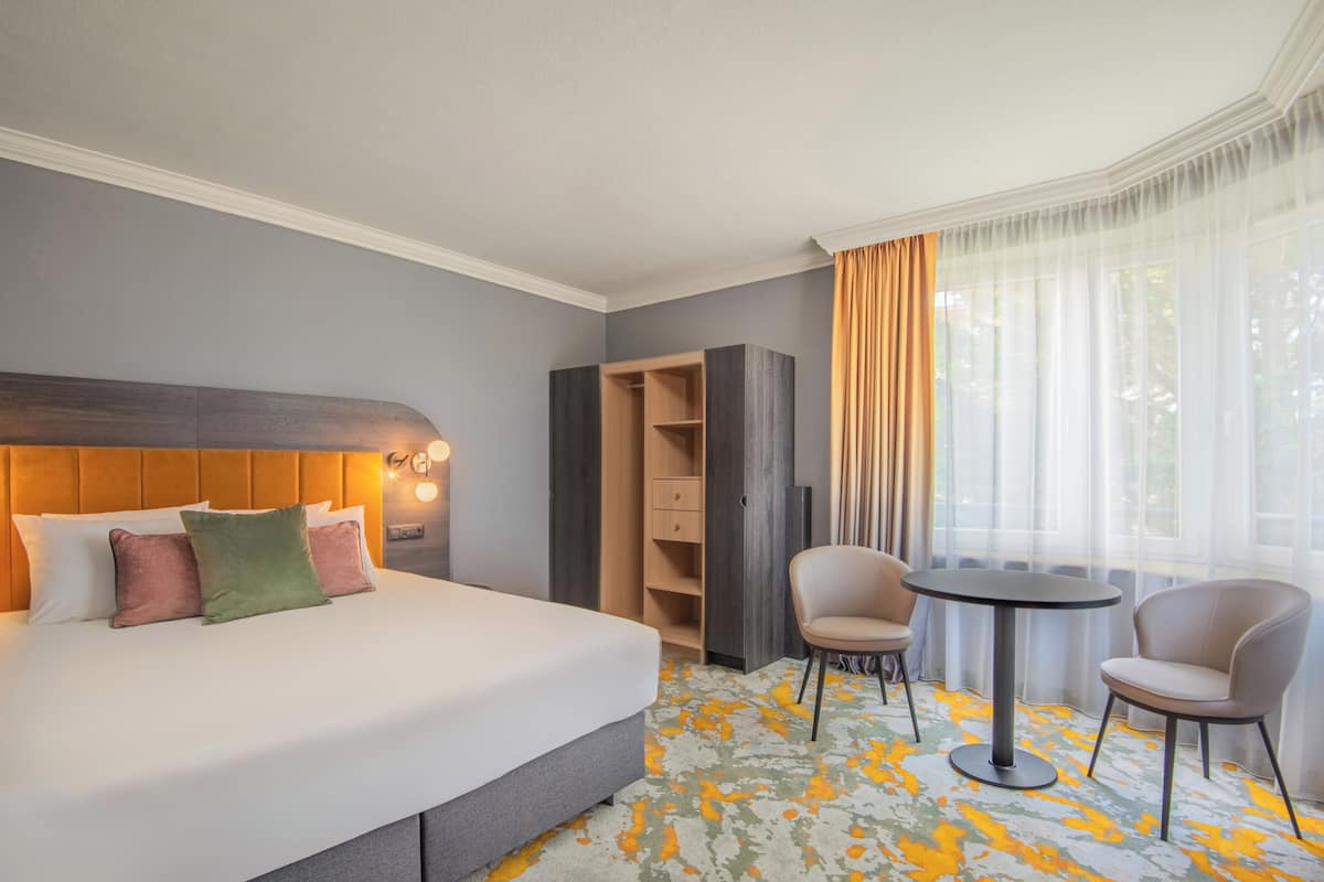 A hotel room with a bed, a seating area with two chairs, a small table, a wardrobe, and a large window with curtains.