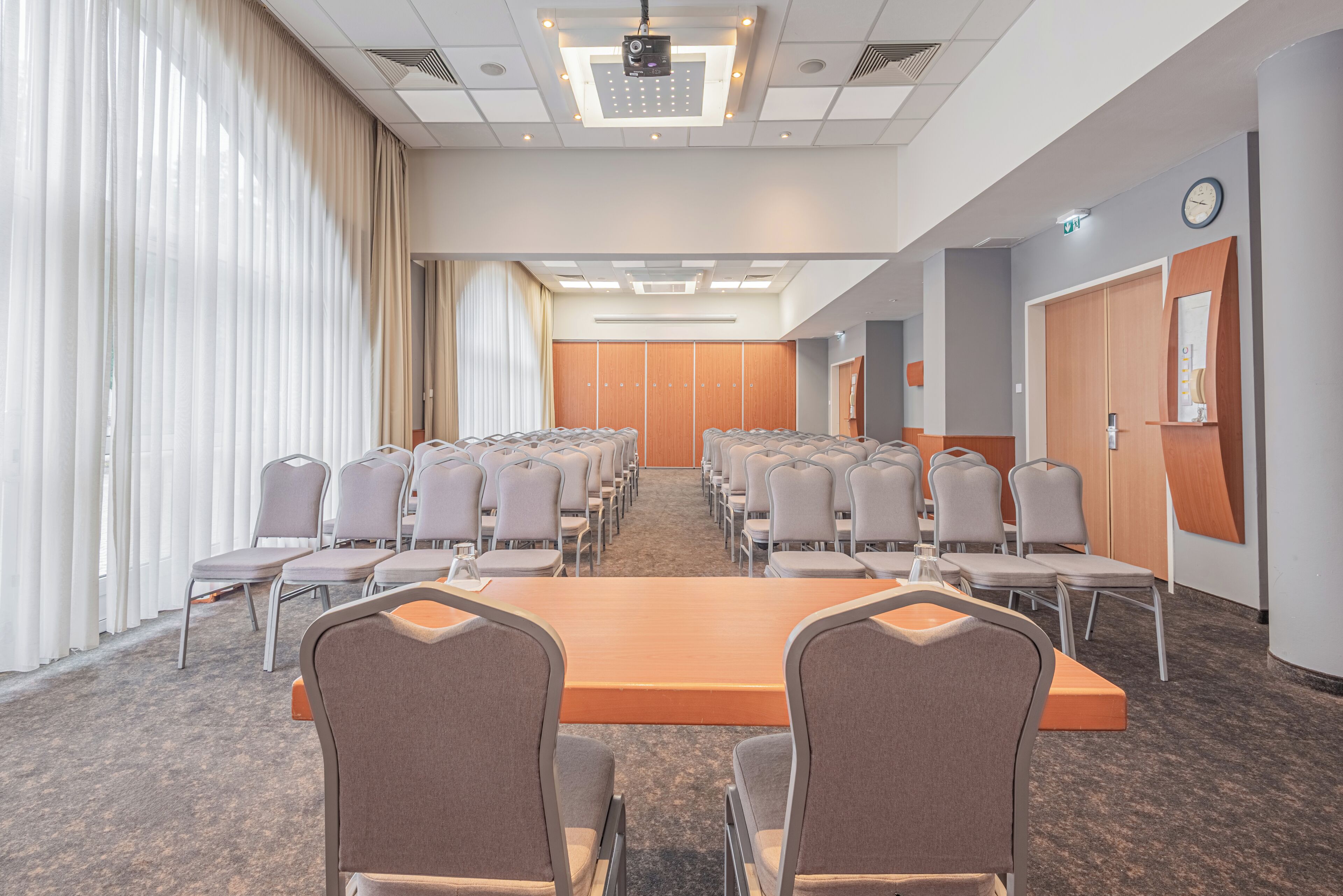 A conference room with a long table and multiple chairs, large windows with curtains, and a clock on the wall.