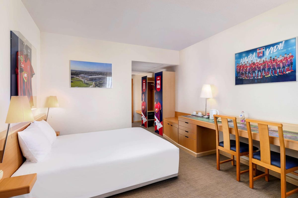 A hotel room with a bed, desk, chairs, a lamp, a poster, and a door leading to a closet.