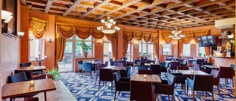 Un restaurante espacioso con techos altos, candelabros y una alfombra estampada. Mesas y sillas dispuestas para comer.