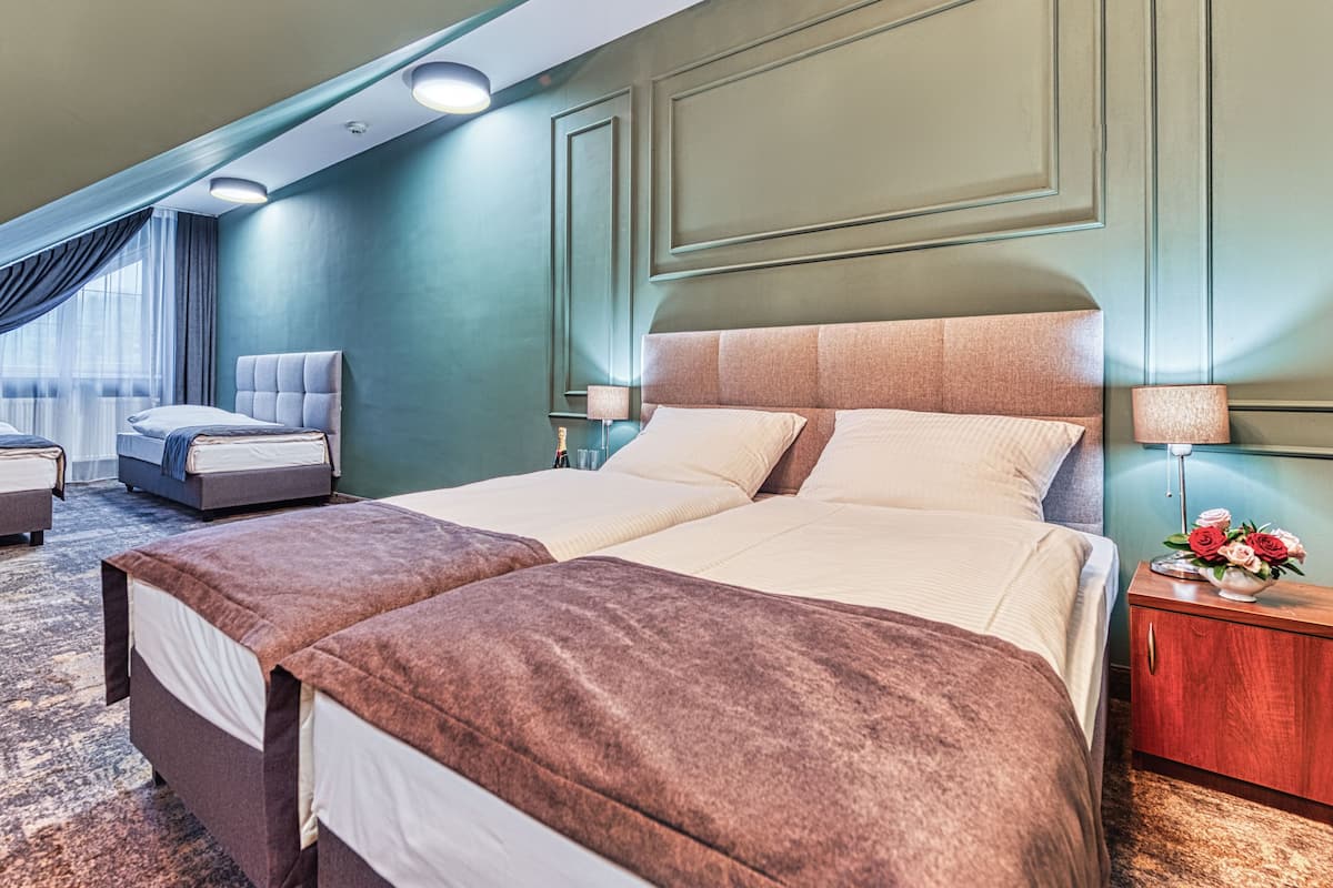 A hotel room with a large bed, a smaller bed, a sofa, a nightstand with a lamp, and a vase with flowers.