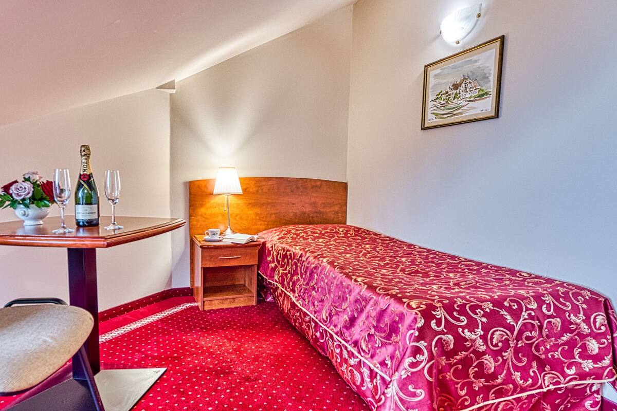 A hotel room with a single bed, a small table with a bottle of champagne and glasses, a chair, a nightstand with a lamp, and a framed picture on the wall.