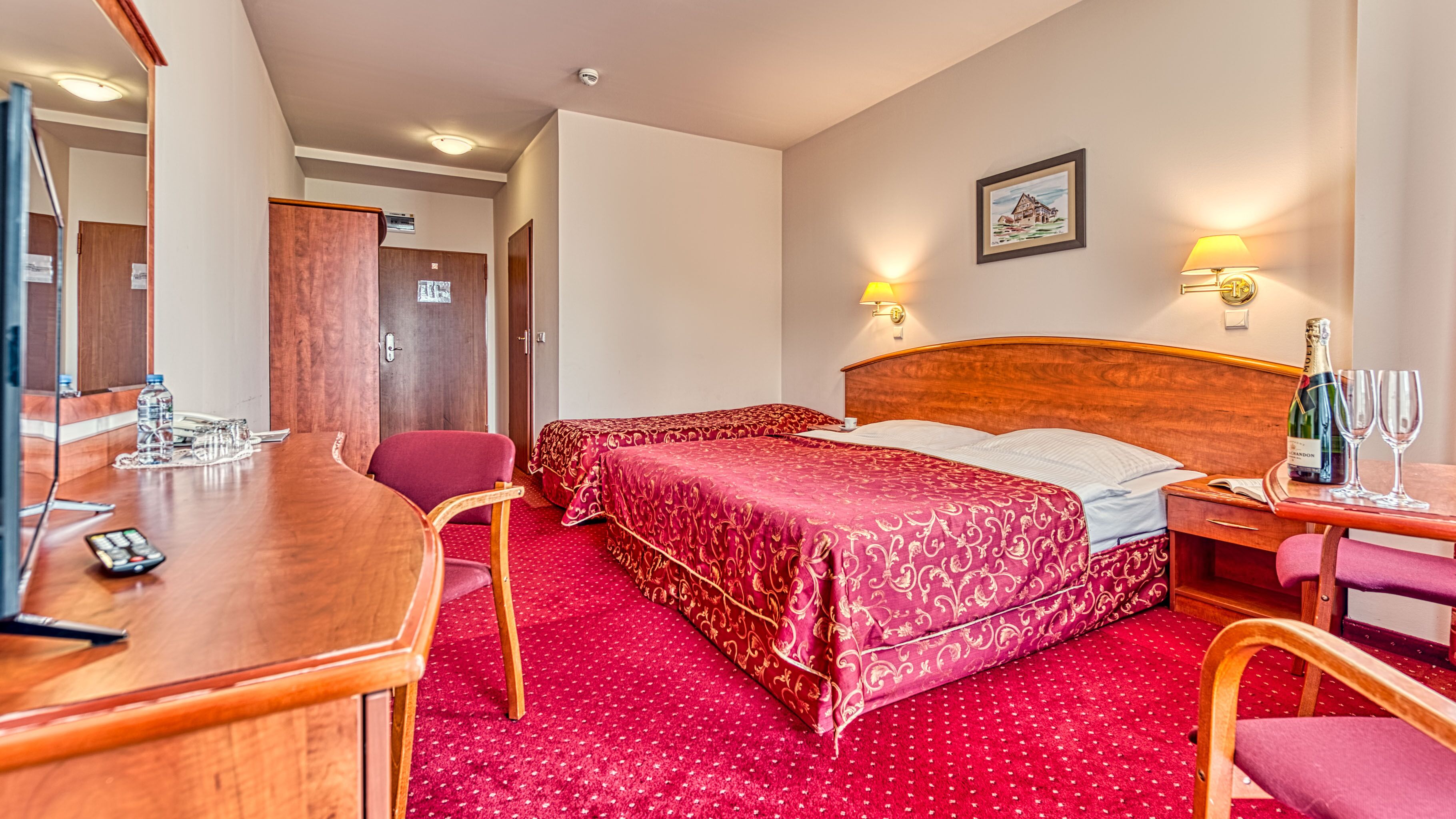 A hotel room with a wooden bed, two bedside tables, a desk with a chair, a mirror, and a red carpet.