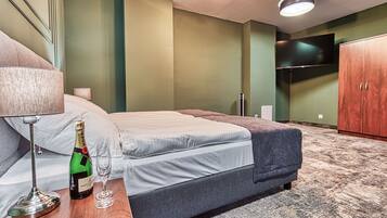 A neatly made bed with a grey blanket, a bedside table with a lamp and a bottle of champagne, and a television mounted on the wall.