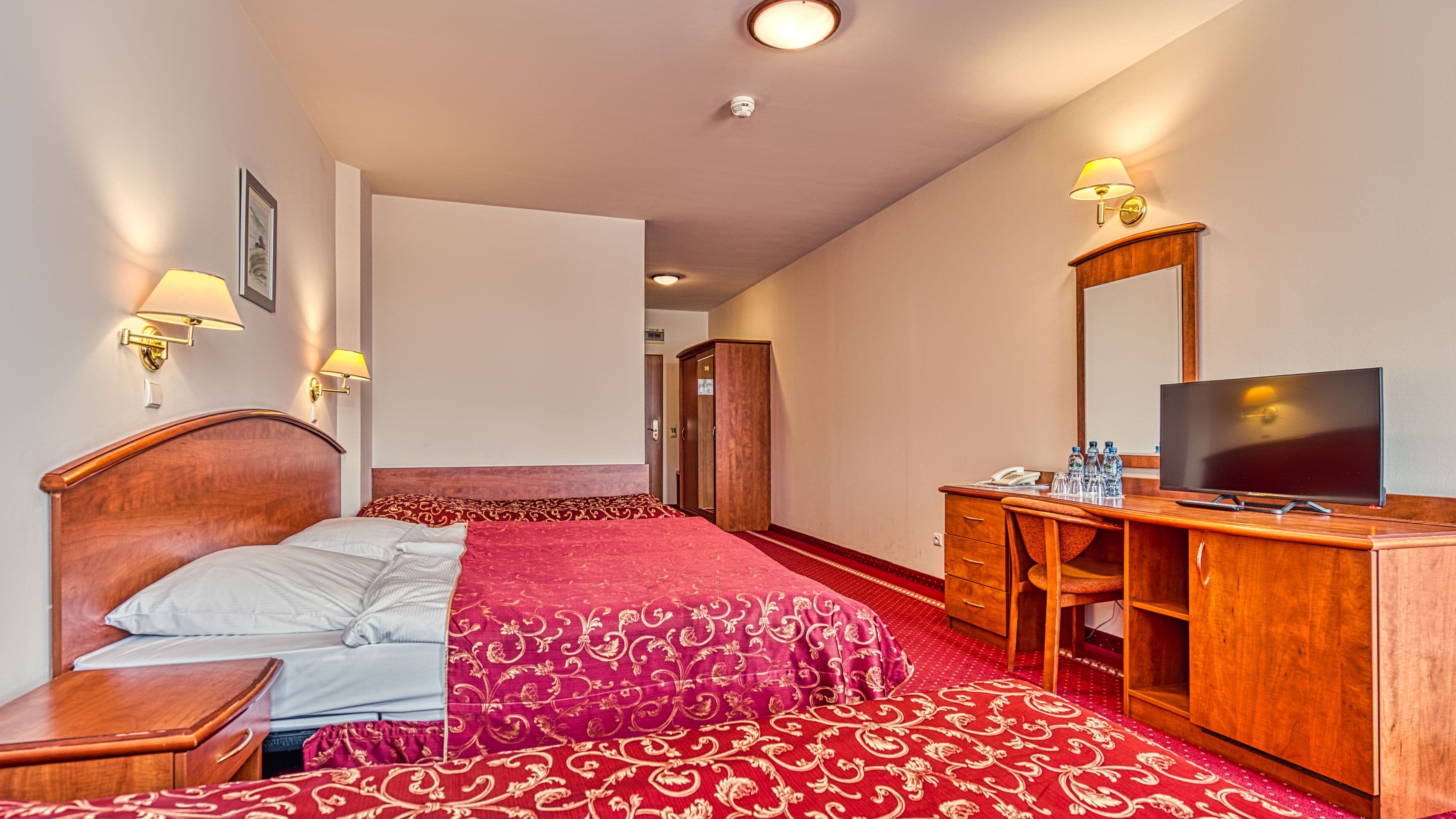 A hotel room with a bed, a wooden dresser, a television, and two wall-mounted lamps.