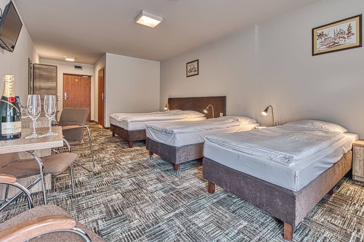 A hotel room with two beds, a desk with a bottle of champagne and glasses, a chair, and a framed picture on the wall.