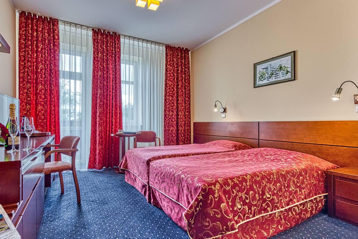 A hotel room with two beds, a desk with a chair, a small table, and a large window with red curtains.