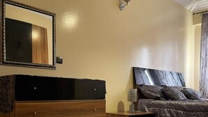 Ein Schlafzimmer mit einem hölzernen Schminktisch, einem Bett mit dunklem Kopfteil, einem Nachttisch, einem Spiegel und einem roten Teppich.