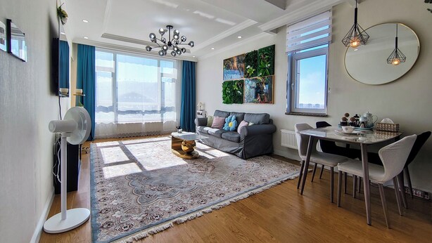 Un salon moderne comprenant un canapé, une table basse, une table à manger et des chaises. Il y a une grande fenêtre offrant une belle vue, un lustre et un ventilateur de plafond.