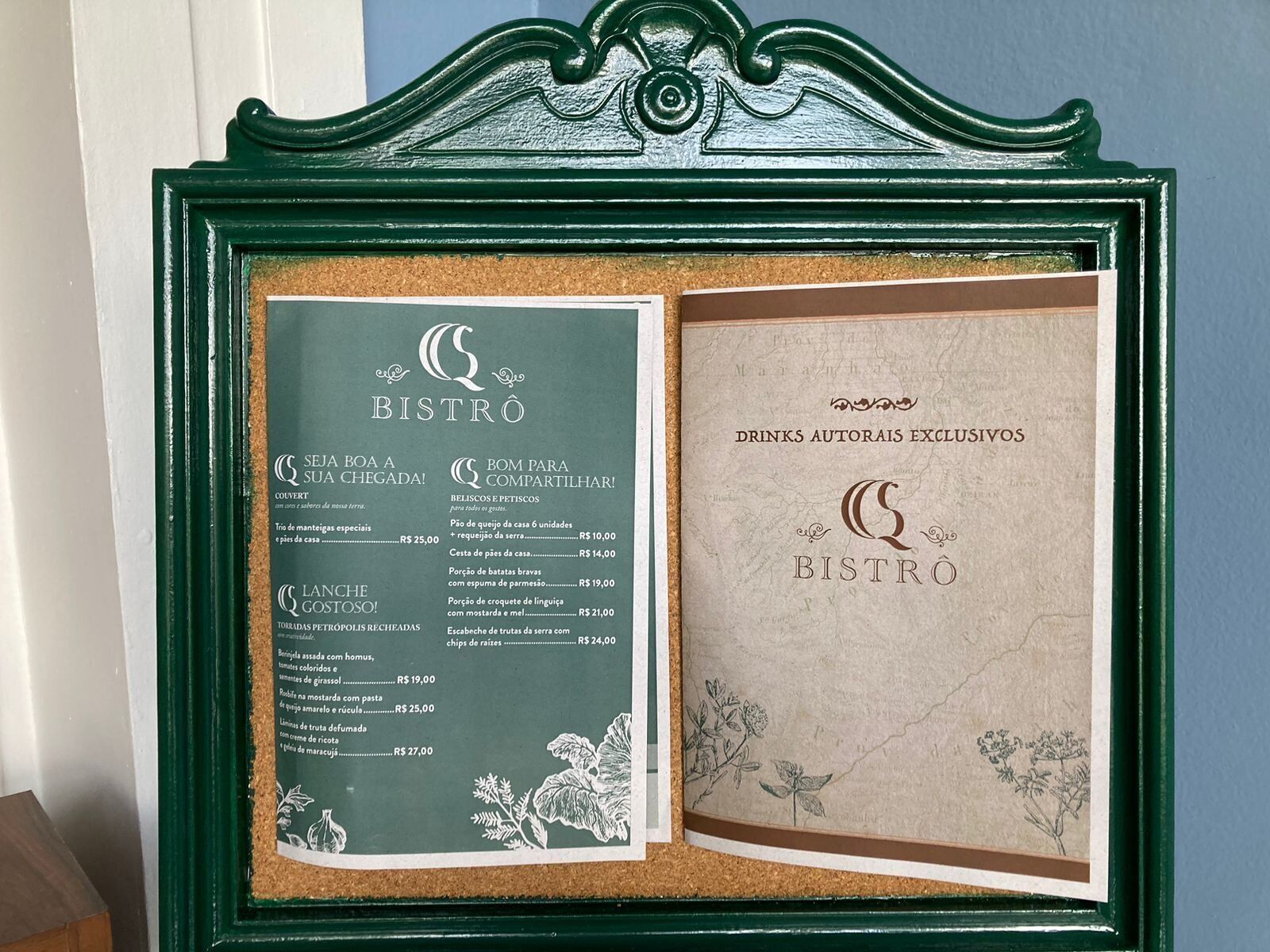 A corkboard menu board with a green frame, listing various food and drink items with prices.