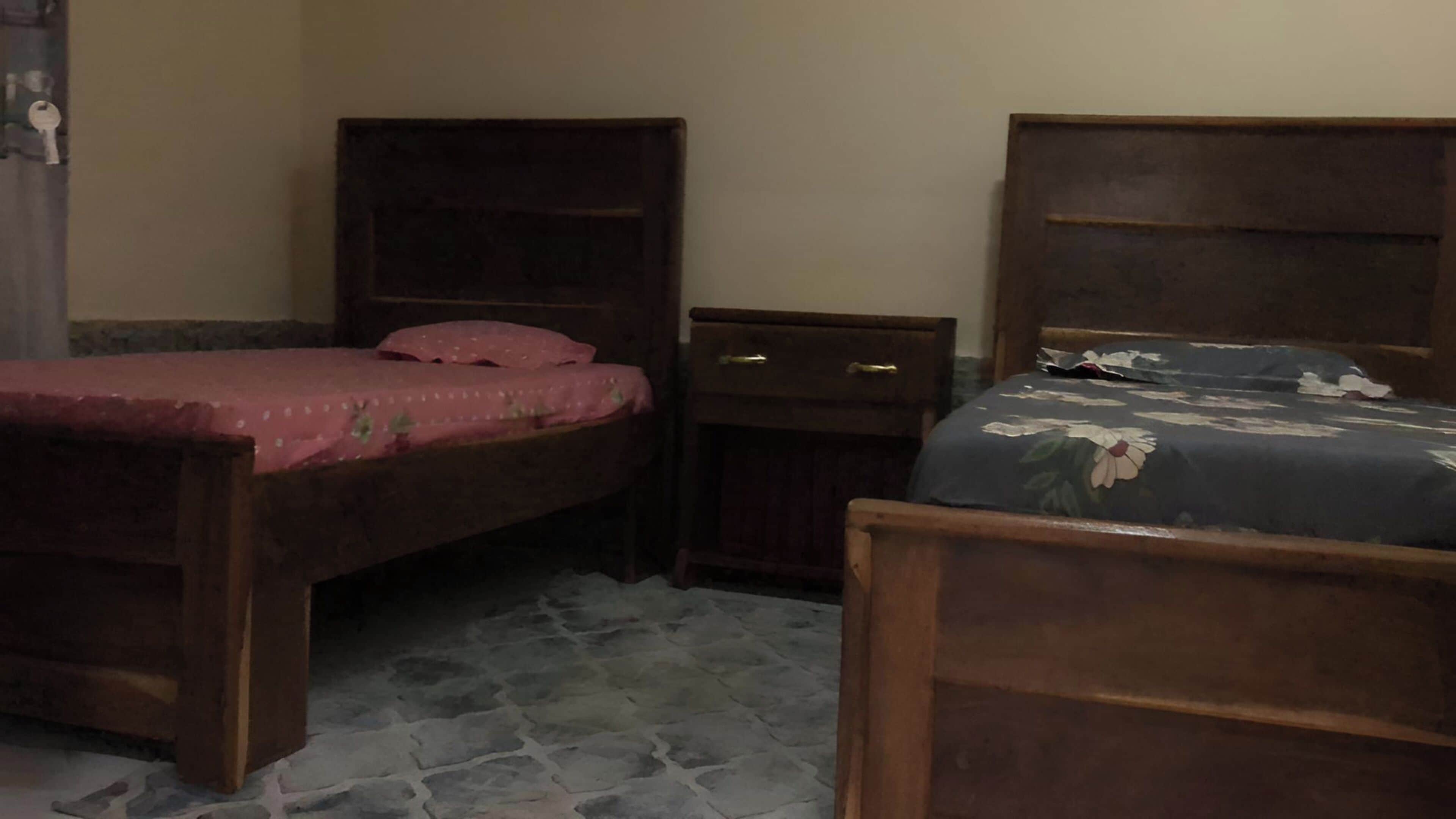 A bedroom with two wooden beds, one with a pink bedspread and the other with a floral bedspread. There is a small wooden nightstand between the beds.