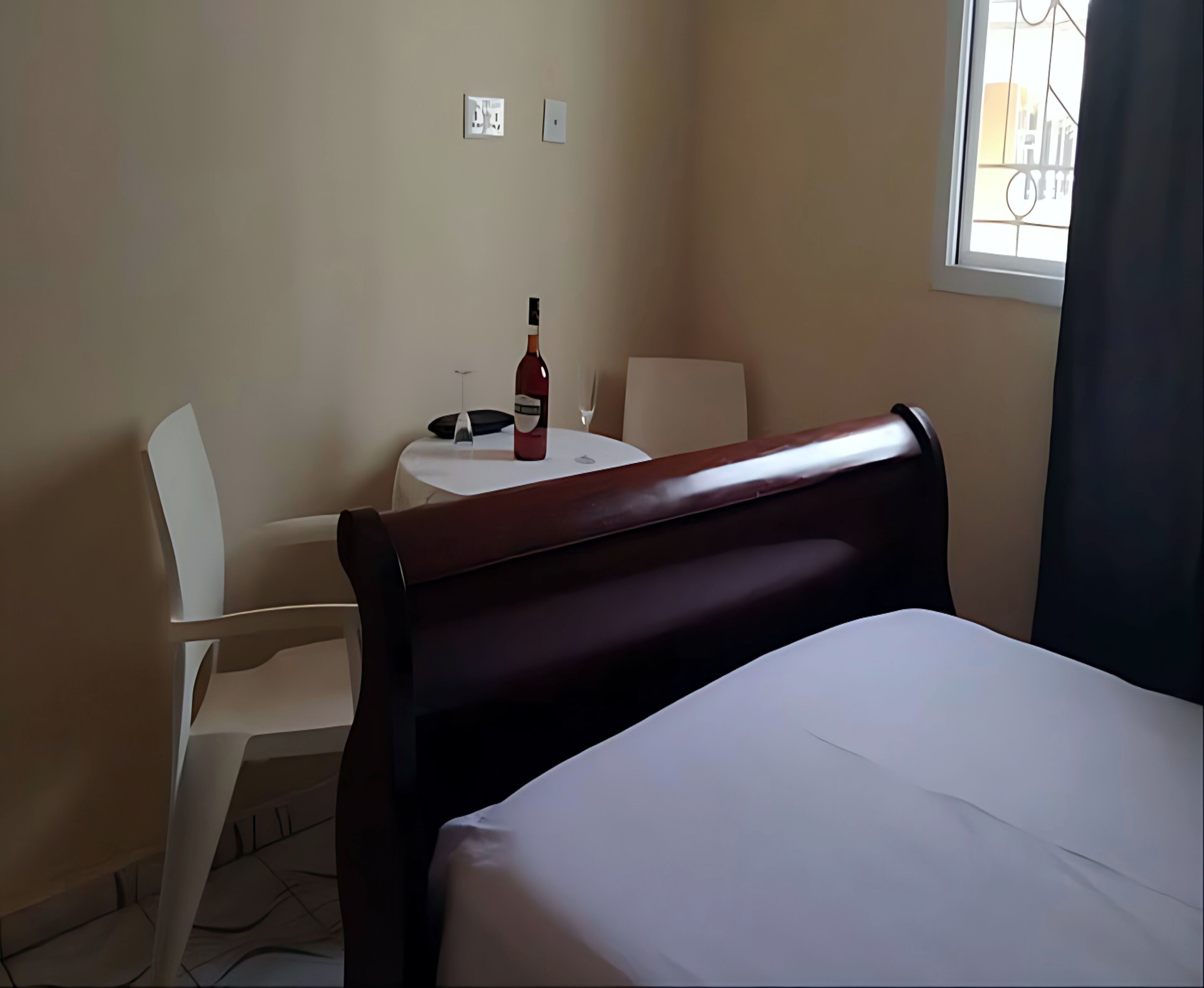 A hotel room with a bed, a small table with a wine bottle and glass, a chair, and a window with a view.