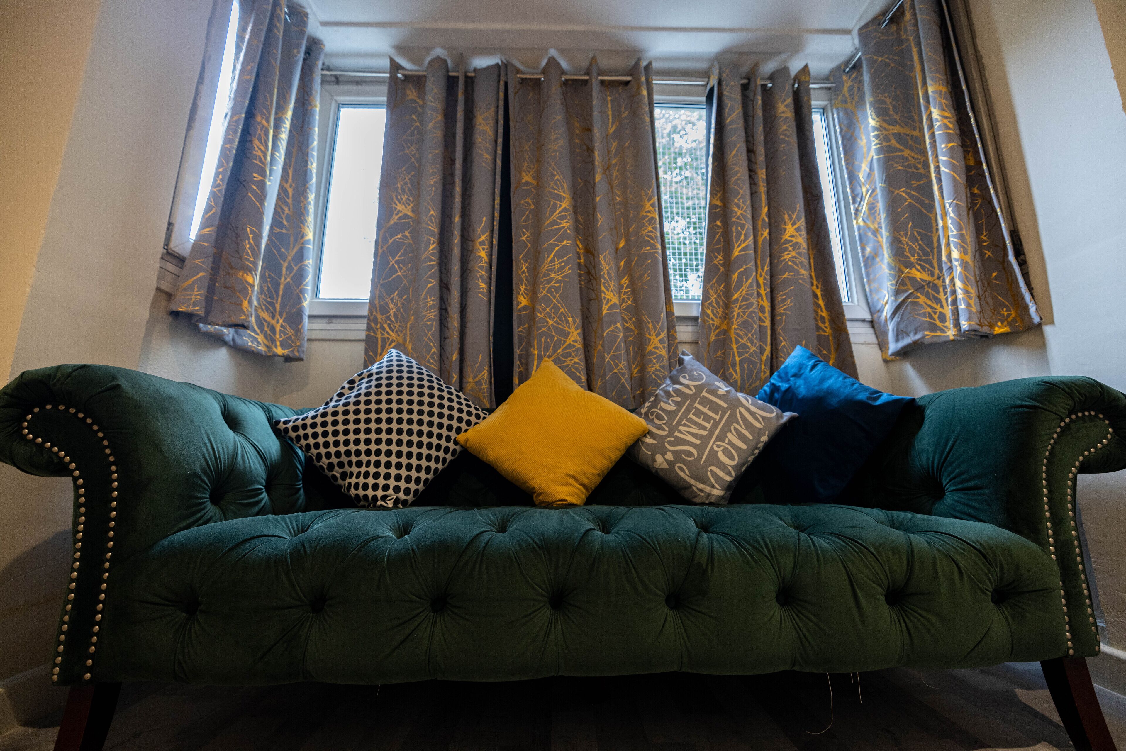 A green tufted sofa with multiple patterned cushions in a room with a window and curtains.