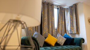 Um sofá verde com várias almofadas, um candeeiro de chão e cortinas com padrão de árvores.