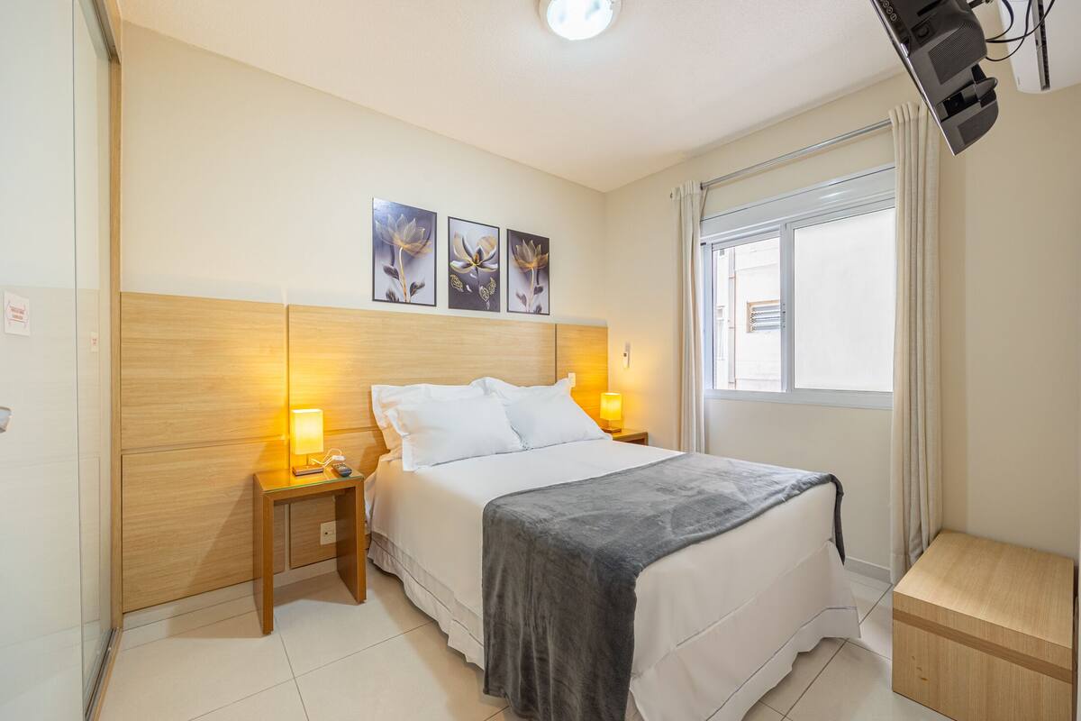 Un dormitorio con cama, lámparas de noche, cabecera de madera, ventana con cortinas y una obra de arte en la pared.