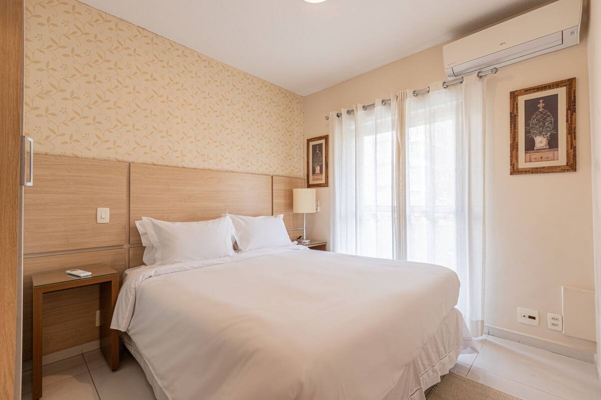 Habitación de hotel con una cama grande, una mesita de noche, una lámpara, un marco de fotos y una ventana con cortinas.