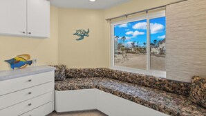 Una habitación con un banco curvo, un armario blanco, un mural en la pared de una tortuga marina y una ventana que ofrece vistas a una playa y casas.