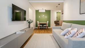 A modern living room with a sofa, a TV mounted on the wall, a dining table with chairs, and a kitchen area in the background. - WI-FI 180MB | 200m from Beiramar Shopping #CA52 (Florianópolis)