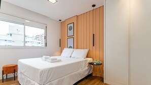 A modern bedroom with a large bed, wooden panel walls, a window with a city view, and a small side table with a book. - WI-FI 180MB | 200m from Beiramar Shopping #CA52 (Florianópolis)