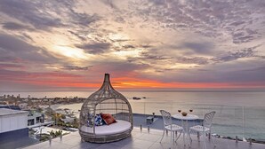 A rooftop terrace with a hanging chair, a table with chairs, and a view of the sea at sunset.