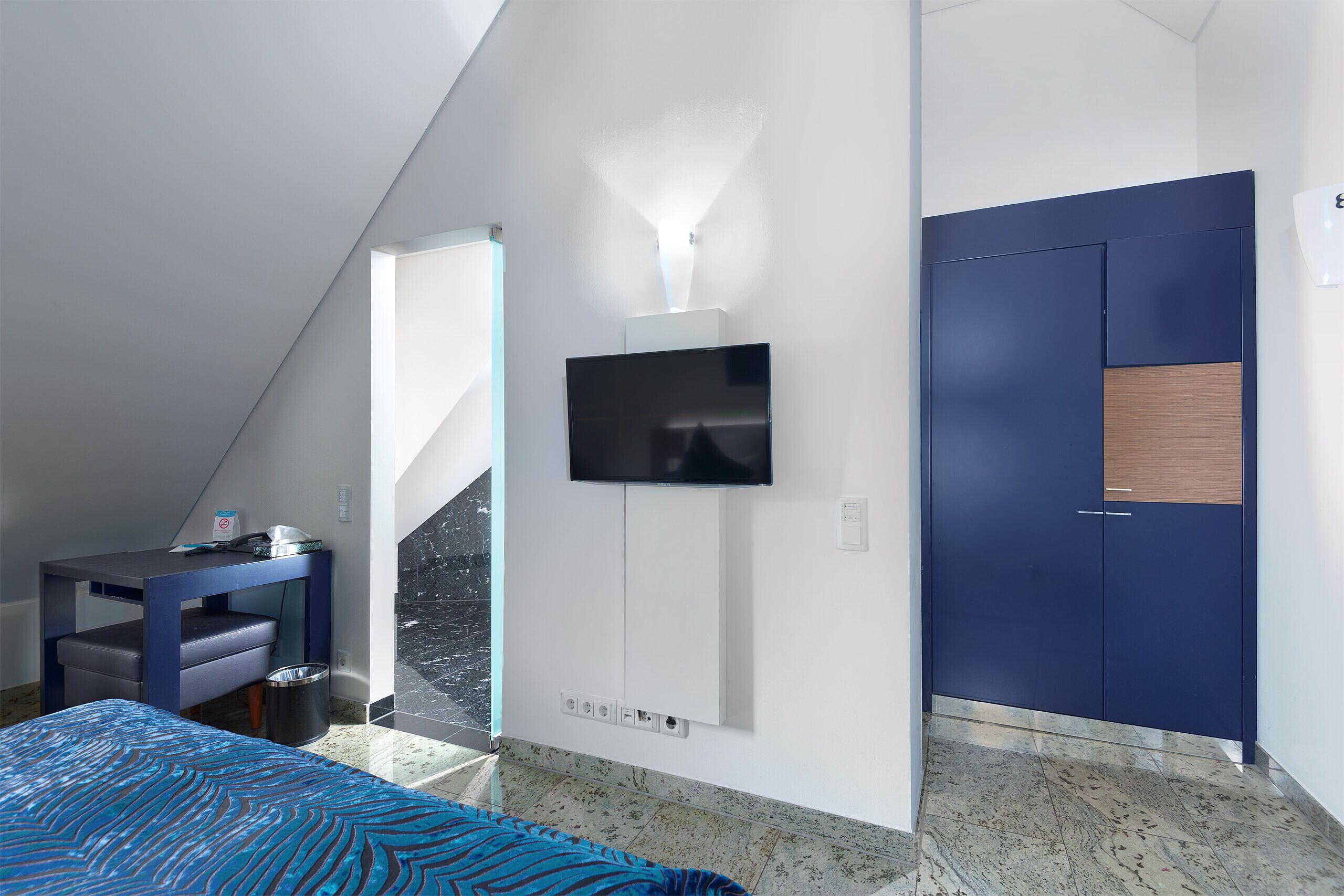Una habitación de hotel moderna con una cama, un escritorio, un televisor montado en la pared y un armario.