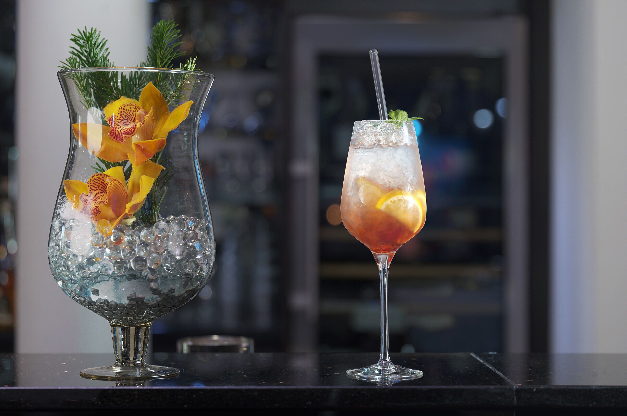 Dos bebidas sobre una barra, una en una jarra de vidrio y otra en una copa de martini, con hielo y adornos.