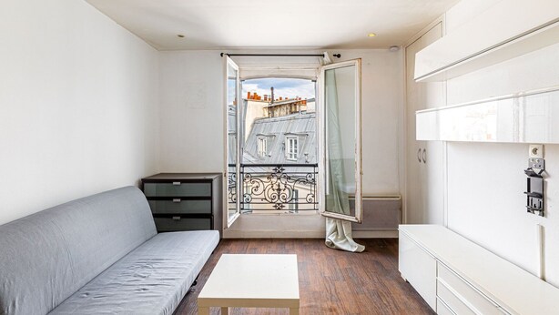 Ein kleiner, moderner Raum mit einem grauen Sofa, einem Holztisch und Blick auf Gebäude durch eine offene Tür.