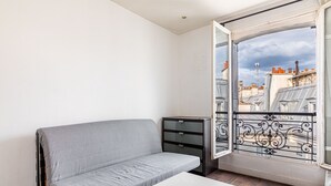 Ein kleiner, moderner Raum mit einem grauen Sofa, einem weißen Tisch und Blick auf eine Stadtansicht durch einen Balkon mit aufwendiger Eisenbalkonage.