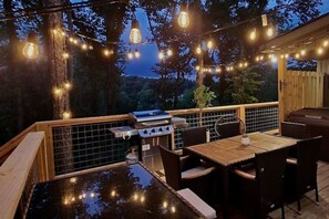 An outdoor patio with a grill, dining table, and chairs, surrounded by trees and string lights. - Mandrake Lodge (Kodak)