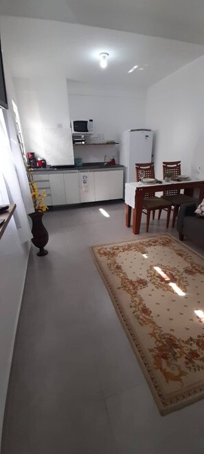 Una cocina moderna con armarios blancos, un microondas y un refrigerador. Hay un comedor con una mesa y sillas, y una alfombra decorativa en el suelo.