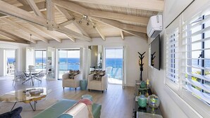 Amplia sala de estar con techo alto, ventanas grandes con persianas y vista al mar.