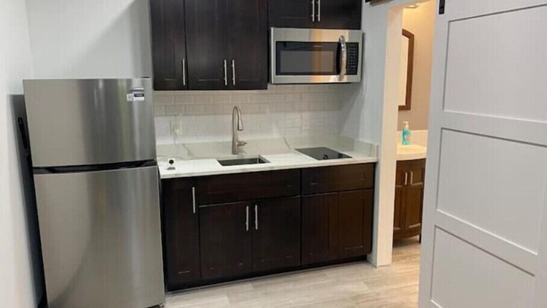 A compact kitchen with dark cabinets, a stainless steel refrigerator, a microwave, and a sink. - Backyard Escape

 (Tampa)