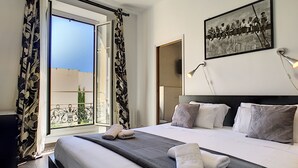 A hotel room with a large bed, a balcony with a view, a painting on the wall, and a bedside lamp.