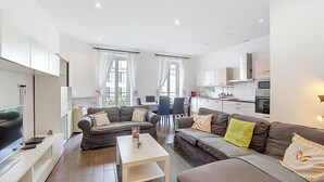 A modern living room with a sofa set, a coffee table, and a TV. There is a dining area with chairs and a table. The kitchen is visible with cabinets and appliances.