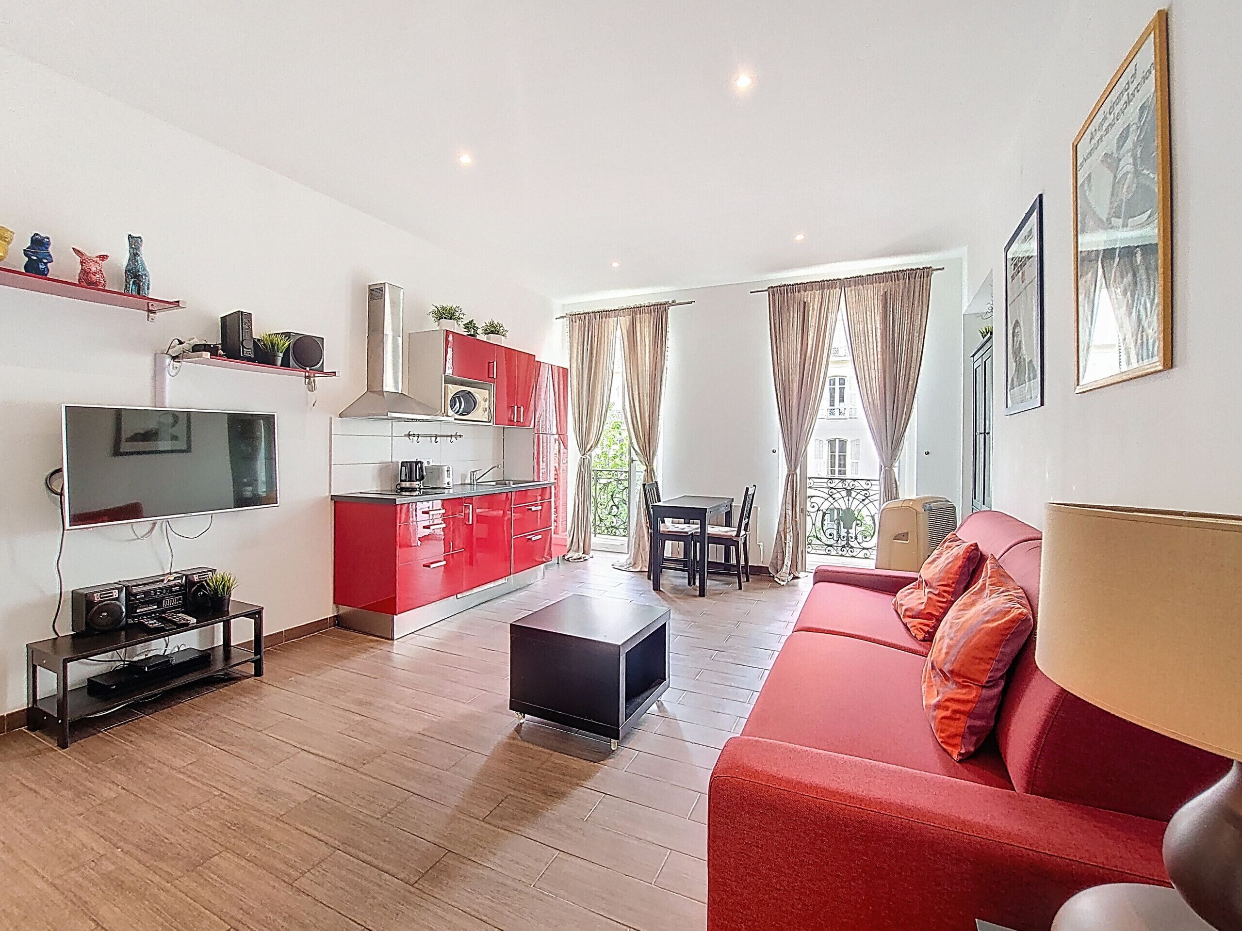 A modern living room with a red sofa, a TV, a kitchen with red cabinets, and a balcony with a table and chairs.