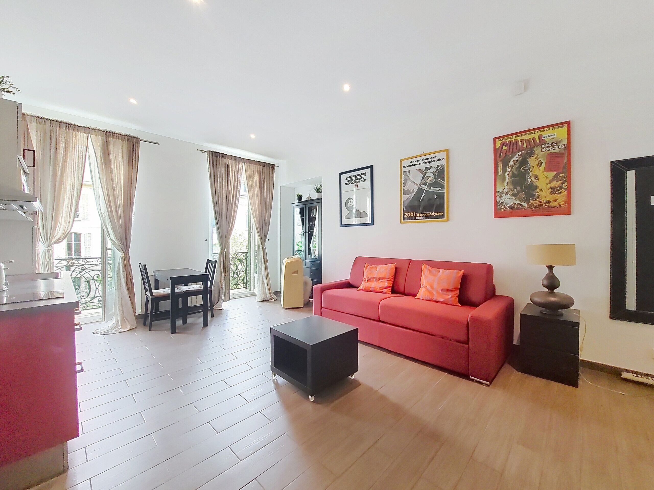 A living room with a red sofa, a black ottoman, a dining table with chairs, and a kitchen area with a sink and cabinets.