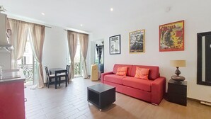 Un salon avec un canapé rouge, un pouf noir, une table à manger avec des chaises et un coin cuisine comprenant un évier et des placards.