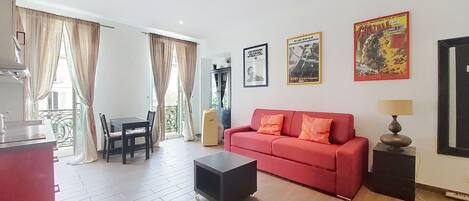 A living room with a red sofa, a black ottoman, a dining table with chairs, and a kitchen area with a sink and cabinets.