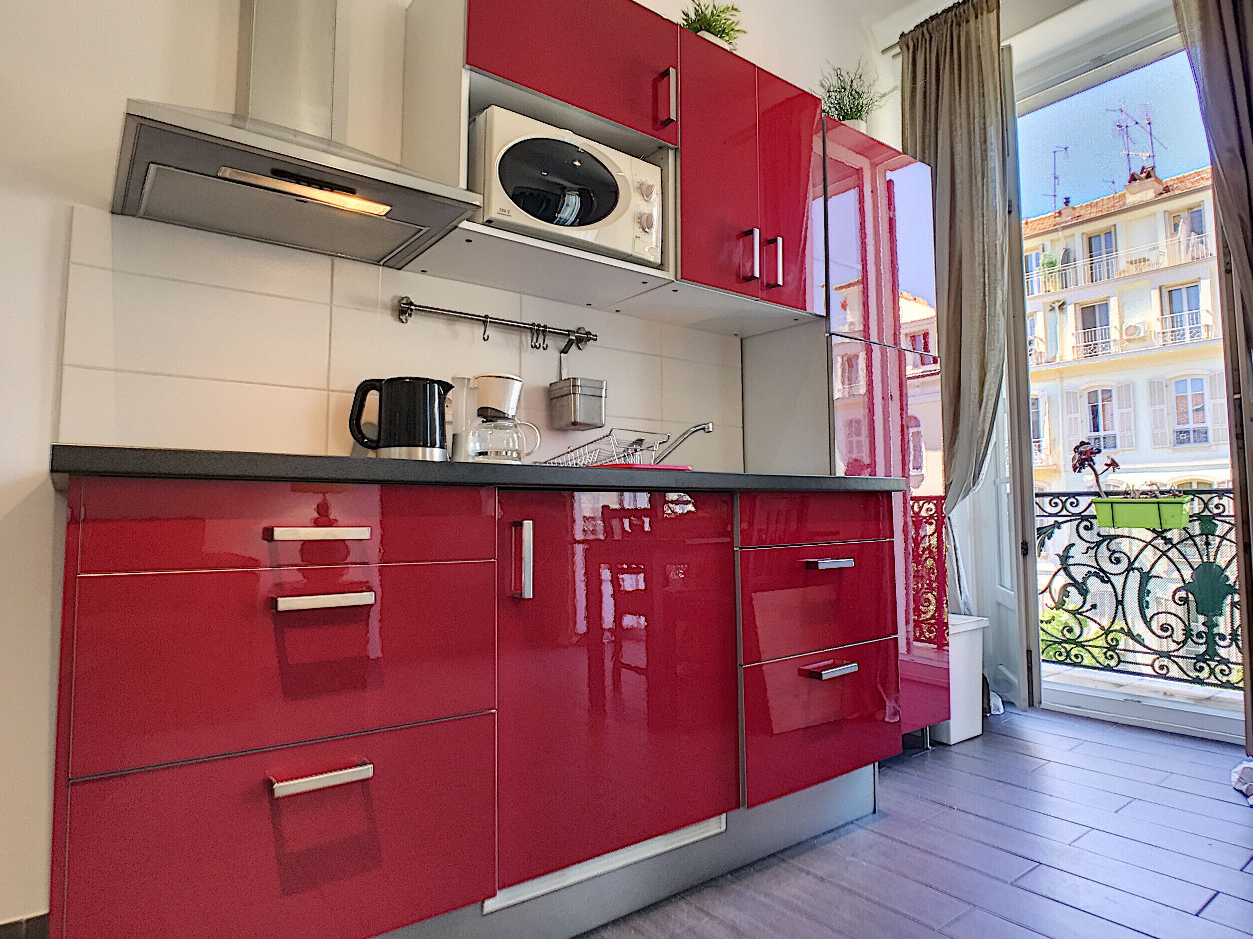 A modern kitchen with red cabinets, a stainless steel range hood, and a view of a balcony with a railing and potted plants.