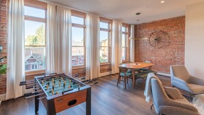 Une pièce avec une table de baby-foot, une table à manger avec des chaises, de grandes fenêtres avec des rideaux, un mur en briques et un luminaire suspendu.