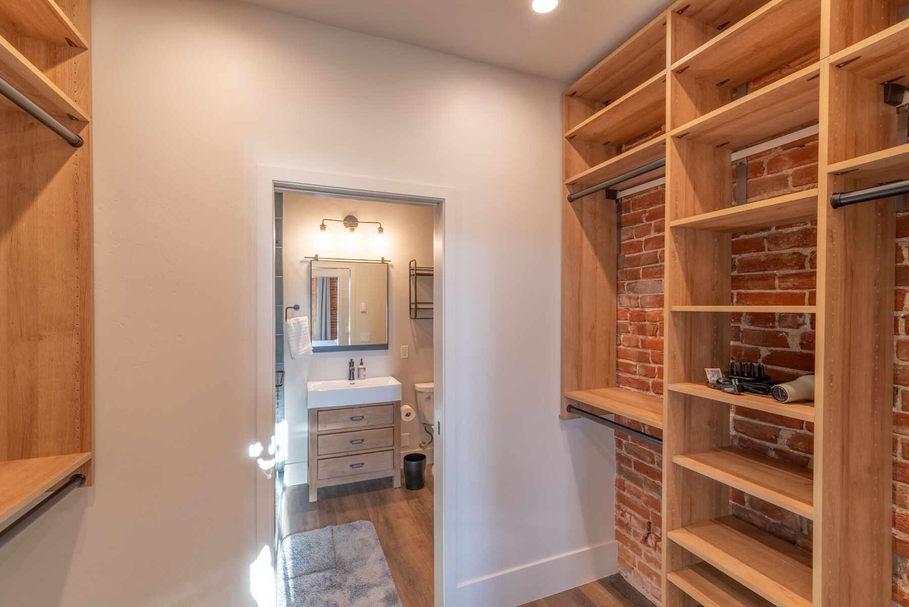 A narrow hallway with a bathroom on the left, featuring a sink, mirror, and towel rack. The right side has a brick wall with wooden shelves.