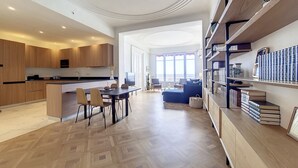 A modern kitchen with wooden cabinets, a dining area with chairs, a sofa, and a bookshelf with books and decorative items.