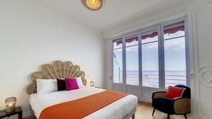 A bedroom with a large bed, a view of the sea through a window, a chair, and a nightstand with a lamp.