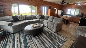 A modern log cabin with a grey sectional sofa, a small ottoman, and a kitchen area. - The Upper Peninsula's Hidden Gem (Rapid River)