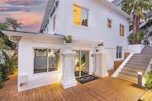 Una casa blanca de dos pisos con terraza de madera, ventanas amplias y una escalera que lleva al piso superior.