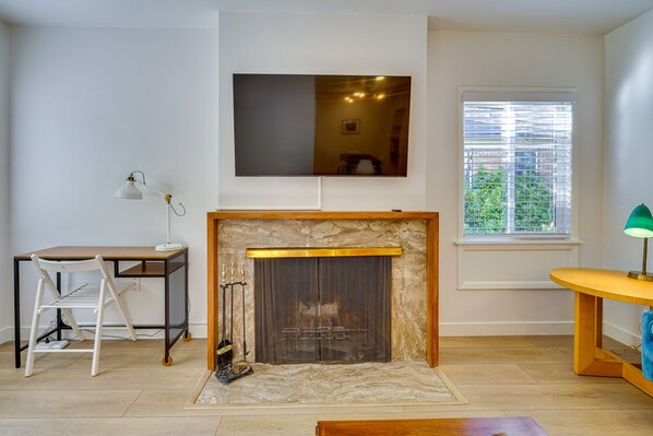 A living room with a fireplace, a TV mounted on the wall, a desk, and a window with blinds.