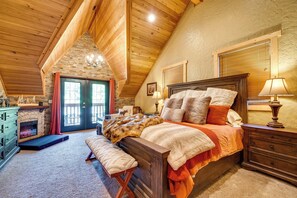 Quarto aconchegante com teto de madeira, lareira de pedra, cama com várias almofadas e um banco.